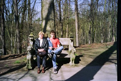 Two individuals sit on a stone bench in a forested park, surrounded by bare trees and dappled sunlight. The woman wears a dar...