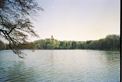 Medieval castle perched on a forested hill, reflected in calm lake waters. Lush greenery surrounds the structure, framed by a...
