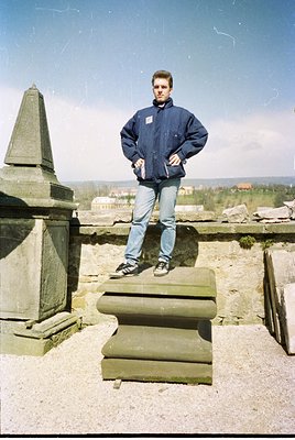 Man in 1980s-style navy jacket and jeans poses atop weathered stone ruins with urban skyline in background. Classic compositi...