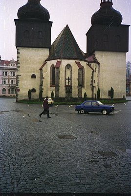 Historic Eastern European church with twin black towers and red-tiled roof, likely 1960s–70s. Cobblestone plaza features a pa...