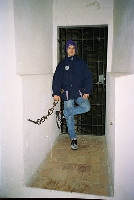 A young individual stands in a confined, windowed alcove with a barred door behind them. They wear a navy puffer jacket, ligh...