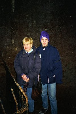 Two individuals pose indoors beside a weathered stone wall, likely part of a historic or industrial site. The man wears a nav...