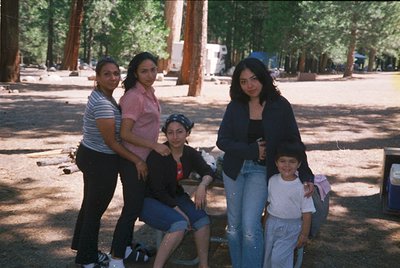 Four individuals pose outdoors in a wooded park setting, likely mid-1990s. Three adults and one child stand/kneel on a dirt p...