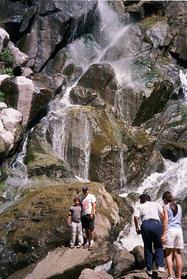 Three adults and a child stand near a cascading waterfall in a rocky mountain setting. The waterfall descends over moss-cover...