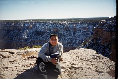 Person kneeling on Grand Canyon rim, holding a camera, dressed in 1990s-style fleece and dark pants. Striking layered red roc...