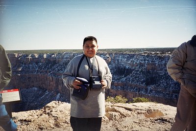 Person in late 20th-century outdoor wear poses with a vintage film camera at Grand Canyon rim, holding a small notebook. Laye...