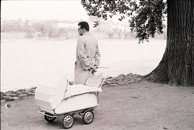 Mid-20th century man in light jacket pushes vintage stroller near waterfront, likely urban park. Classic 1950s–60s attire and...