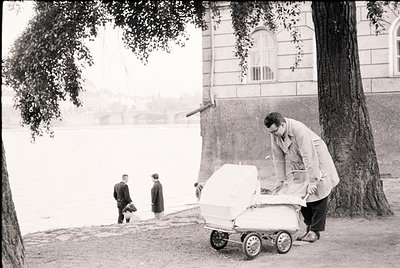 Mid-20th century black-and-white street scene featuring a man in a light coat pushing a vintage baby stroller near a grand, c...