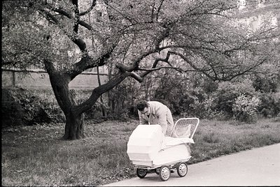 Mid-20th century black-and-white street scene: man in suit adjusts vintage white stroller on paved road beside leafy tree. Ur...