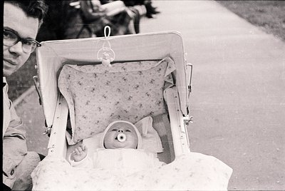A man in mid-20th-century attire holds an open suitcase repurposed as a portable bassinet, containing a sleeping infant wrapp...