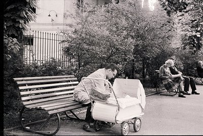 Mid-20th century urban park scene: man in light jacket adjusts vintage stroller beside wooden bench. Elderly woman in plaid s...