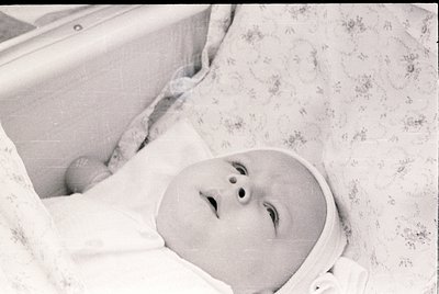 Vintage black-and-white portrait of an infant in a crib with floral-patterned linen, mid-20th century. Classic baby cap and d...