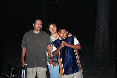 Three individuals pose outdoors at night under dim lighting, likely flashlit. The man wears a gray T-shirt and beige pants; t...