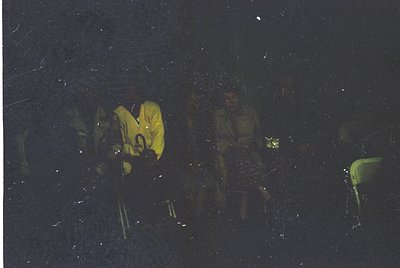 Three figures in camouflage gear and yellow jackets stand on a snowy slope under night sky, illuminated by a single light sou...