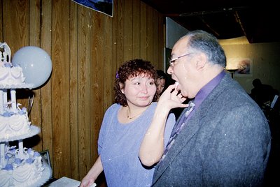 Vintage indoor celebration featuring a multi-tiered wedding cake with floral decorations. A woman in a purple dress and man i...