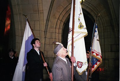 Veterans in formal attire holding flags under a stone archway, likely at a WWII commemoration. The central figure wears a 193...