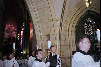 Gothic cathedral interior with stained-glass windows casting vibrant light. Clergy in white vestments process with lit candle...