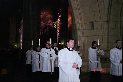 Religious procession inside a Gothic-style church, likely 1990s. Clergy in white vestments carry candles and a crucifix, fram...