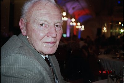 Portrait of an elderly man in a formal suit, captured in low-light indoor setting. His expression is neutral, with a slight s...