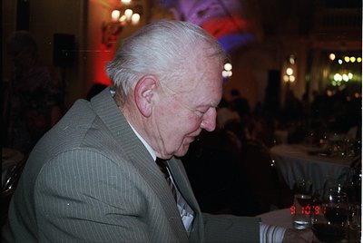 Elegant older gentleman in formal attire—beige blazer, white shirt, and tie—sitting at a dimly lit indoor event. Neon "9:10" ...