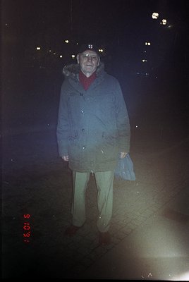 Portrait of an elderly man in winter attire—hooded parka, scarf, and beige trousers—standing on a wet urban street at night. ...