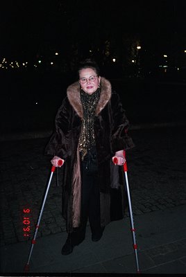 Portrait of an elderly woman in a winter coat with fur collar, standing outdoors at night. She wears glasses and holds white ...