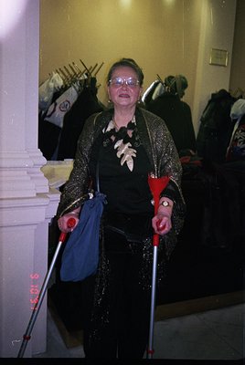 Elegant woman in 1960s-70s formalwear—sparkling sequined jacket, black dress, and red cane—poses indoors near a staircase. De...