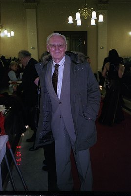 Mid-century formal event in a grand hall, featuring a man in a dark blazer, striped tie, and glasses. Ornate chandeliers and ...