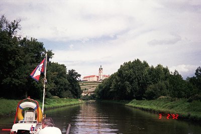A vintage boat cruises a calm river flanked by lush greenery, with a castle-like complex in the background. The structure fea...