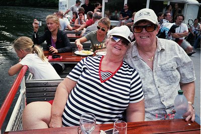Two adults pose joyfully on a covered canal boat, likely a 1990s Dutch *taxi boot* (boat taxi). The man wears a light cap and...