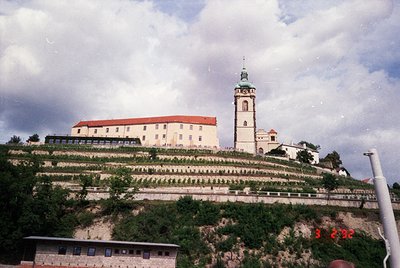 Historic hilltop monastery with red-tiled roof and central bell tower, surrounded by terraced greenery. Likely Eastern Europe...