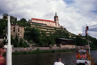 Historic castle perched on a hillside overlooking a river, featuring red-tiled roofs and a prominent tower. Mid-20th century ...