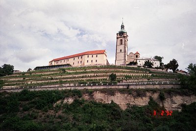 Historic monastery complex perched on terraced hillside, featuring a prominent bell tower with green dome and red-tiled roofs...