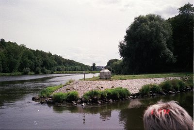 Vintage riverbank scene with concrete steps leading into calm waters, bordered by grassy embankment and dense foliage. Overca...