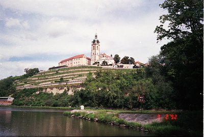 Historic hilltop fortress with red-tiled roofs and a central bell tower, surrounded by terraced greenery. Overlooking a calm ...