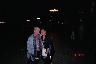 Two women pose under illuminated streetlights in an urban plaza, likely during a nighttime event. The woman on the left wears...