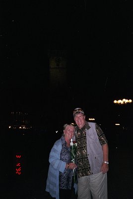 Couple posing under nighttime city lights, holding a single white flower. Man wears patterned shirt, white cap, and light-col...