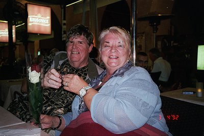 Couple posing indoors at a social event, holding a bouquet of red roses. Man wears patterned shirt, watch, and dark trousers;...
