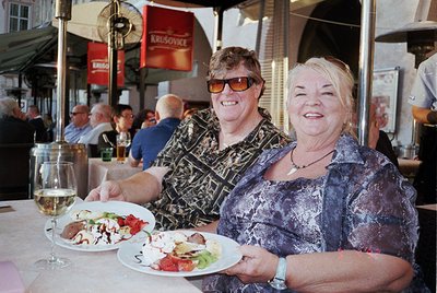 Two women pose joyfully at an outdoor café, holding plates with salads and meats. The woman on the left wears patterned sungl...
