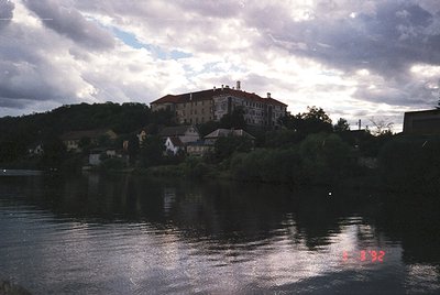 A vintage photograph of a riverside settlement with a prominent multi-story building atop a hill, likely a hotel or instituti...