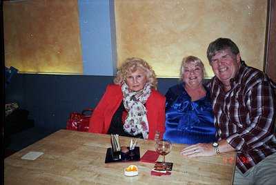 Three adults pose indoors at a wooden table with wine glasses, candlesticks, and a small plate of food. The woman on left wea...