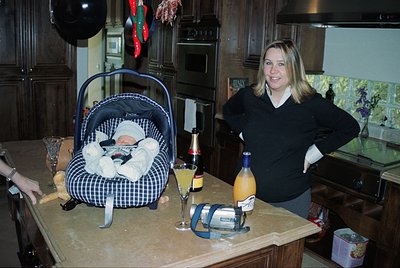 A woman in a black top poses beside a kitchen island with a baby in a blue/gray carrier. Wooden cabinets, a wine bottle, cham...