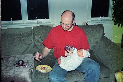 A man in a red shirt holds a baby on a sofa, eating a plate of food while holding a remote. Indoor setting with a potted plan...