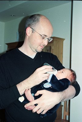 A man in a black V-neck shirt cradles an infant in a dark onesie, feeding with a bottle. Indoor setting with wooden furniture...