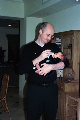 A man in a black sweater cradles an infant in a modern indoor setting, likely a kitchen or living area. Wooden cabinetry with...