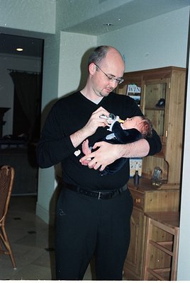 A man in a dark long-sleeve shirt cradles a swaddled infant indoors, likely in a mid-2000s home setting. Wooden cabinetry and...