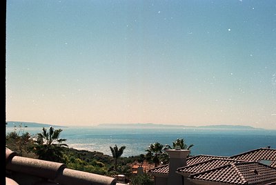 Vintage seaside horizon shot with terracotta rooftops and palm trees in foreground. Clear blue waters meet hazy landmass unde...