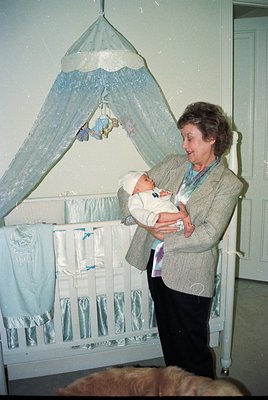 A woman in a 1970s-style cardigan cradles a swaddled infant in a vintage nursery. Decorative canopy bed with blue lace drapes...