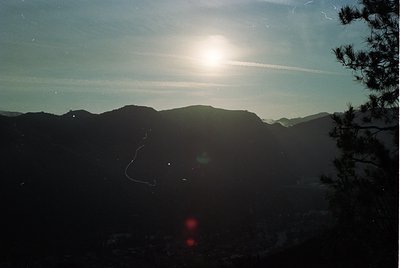 Sunburst through thin clouds illuminates rugged mountain peaks, creating lens flare. Silhouetted pine tree frames right edge....