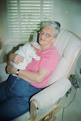 Mid-20th century indoor portrait: Elderly woman in a pink blouse and glasses cradles a swaddled infant in a vintage armchair,...
